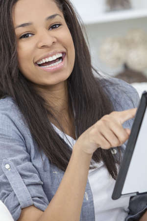 Beautiful happy young African American woman or girl smiling and using a tablet computer at home on her sofaの写真素材