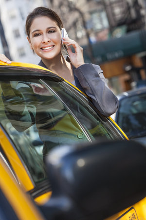 A happy young woman talking on her mobile cell phone by a yellow taxi cab. Shot on location in New...