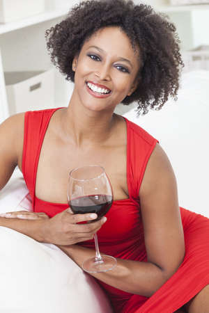 A beautiful happy mixed race African American girl or young woman wearing a red dress and drinking red wine at homeの写真素材