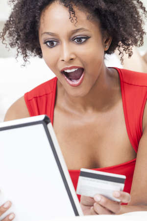 A beautiful, happy and surprised, mixed race African American girl or young woman laying down wearing a red dress using a tablet computer and credit card internet shopping on lineの写真素材