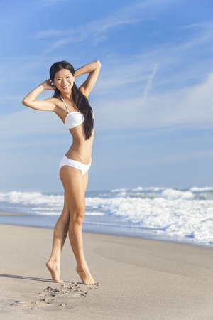 A beautiful Chinese Asian young woman or girl wearing a white bikini on an empty deserted tropical beach with a blue sky の写真素材