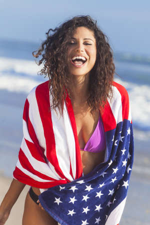 Beautiful young woman laughing wearing bikini and wrapped in American flag towel on a sunny beachの写真素材