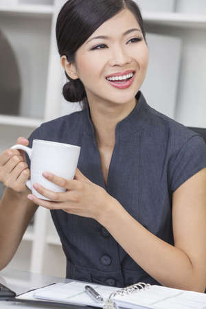 Beautiful young CChinese Asian woman businesswoman smiling, relaxing and drinking a cup of coffee or tea in her officeの写真素材