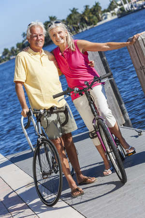 Happy senior man and woman couple together cycling on bicycles with bright clear blue sky by a river or seaの写真素材
