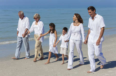 A happy family of grandparents, mother, father and two children, son and daughter, walking holding hands and having fun in the sand of a sunny beachの写真素材