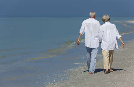 Rear view of happy senior man and woman couple walking and holding hands on a deserted tropical beach with bright clear blue skyの写真素材