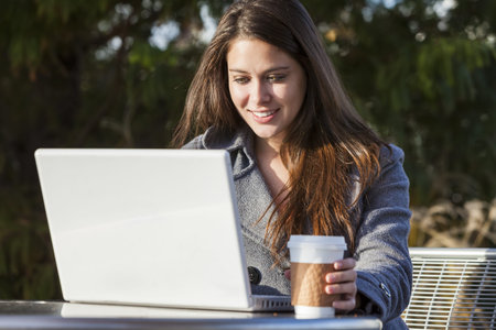 A young woman or girl student using a laptop outside and drinking takeaway coffee