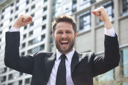 A young successful man, male executive businessman arms raised celebrating cheering shouting in front of a high rise office block in a modern cityの写真素材