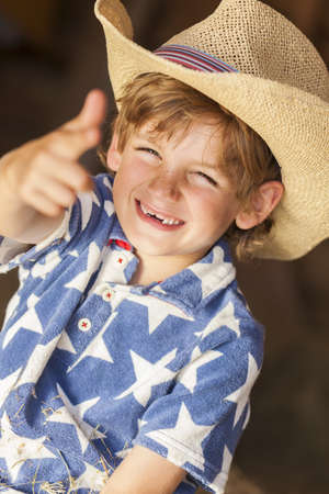 Young happy smiling blond boy child wearing a blue star shirt and cowboy hat sitting on hay or straw bales making a finger gunの写真素材