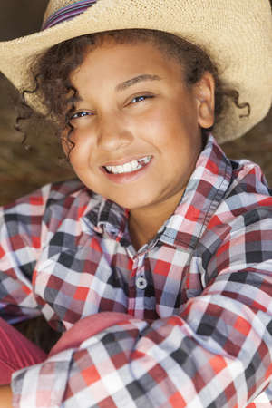 A beautiful and happy mixed race African American female girl child wearing straw cowboy hat and plaid shirt sitting in hay filled barnの写真素材