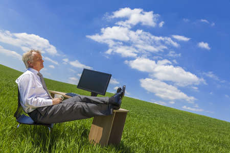 Man or male businessman relaxing feet up at a desk with a computer in a green field drinking tea or coffeeの写真素材