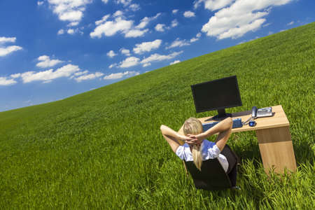 a beautiful young woman or businesswoman relaxing at a desk with computer in a green field with a bright blue sky  の写真素材