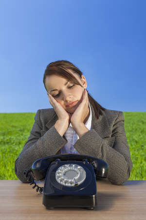 Business concept shot of a beautiful young woman businessowman sitting at a desk waiting for old vintage telephone to call in  green field with a bright blue sky  の写真素材