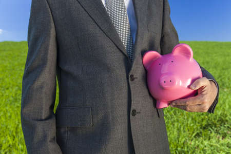 businessman in suit holding a pink piggy bank in a green field with a bright blue sky. の写真素材