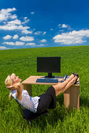 Business concept shot of a beautiful young woman businesswoman relaxing with her feet up at a desk with a computer in a green field with a bright blue skyの写真素材