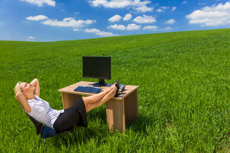 Business concept shot of a beautiful young woman businesswoman relaxing with her feet up at a desk with a computer in a green field with a bright blue skyの写真素材