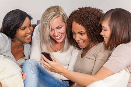 Interracial group of three beautiful young women girl friends at home sitting together on a sofa smiling, surprised and shocked using cell phone smartphoneの写真素材