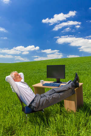 Man or male businessman relaxing feet up at a desk with a computer in a green field drinking tea or coffeeの写真素材