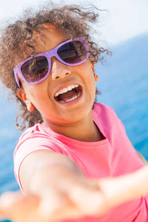 A beautiful and happy mixed race African American female girl child wearing sunglasses in bright sunshine on vacation in front of a blue tropical seaの写真素材