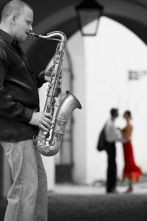 Black and white photograph street musician playing his saxophone while a romantic couple can be seen out of focus in the background the woman is in color wearing a red dressの写真素材