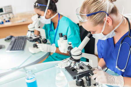 A caucasian female medical or scientific researcher or doctor using her microscope in a laboratory with her Asian Indian colleague beside her.の写真素材