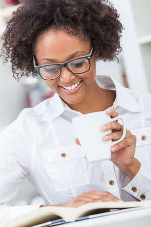 A beautiful happy mixed race African American girl or young woman wearing glasses drinking coffee or tea and reading a book in her kitchen at homeの写真素材