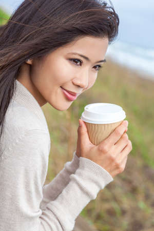 Beautiful young Asian Chinese woman or girl outside, smiling and drinking a takeaway cup of coffee or teaの写真素材