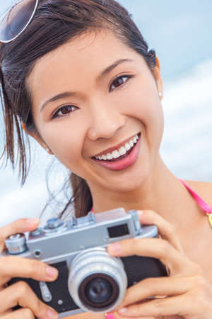 A beautiful Asian Chinese girl or young woman wearing bikini at a beach looking happy taking pictures or photographs with a retro digital cameraの写真素材