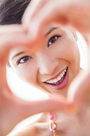 Beautiful young Chinese Asian woman girl smiling, making and looking through a hand heart shaped finger frameの写真素材