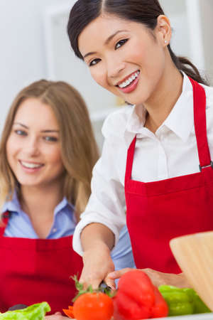 Beautiful happy young Asian Chinese woman or girl and her friend or work colleague wearing a red apron cutting & preparing fresh vegetable salad food in a kitchenの写真素材