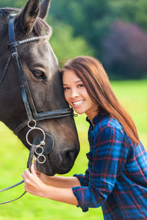 Beautiful happy Asian Eurasian young woman or girl wearing plaid checked shirt, smiling and rsting her head on her horse in sunshineの写真素材