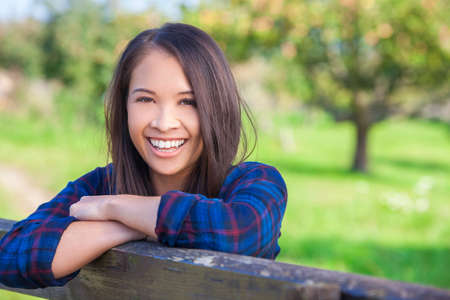 Beautiful happy Asian Eurasian young woman or girl wearing denim shirt, smiling and leaning on fence in sunshineの写真素材