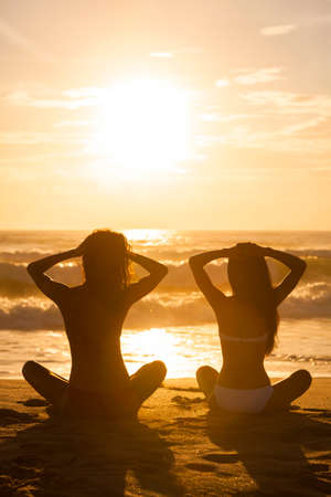 Two relaxed sexy young women or girls wearing bikinis sitting on a deserted tropical beach at sunset or sunriseの写真素材