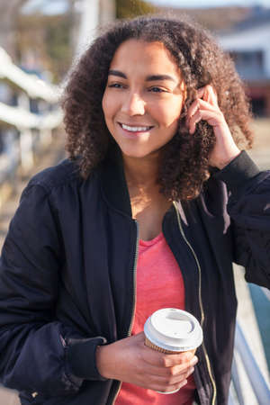 Beautiful happy mixed race African American girl teenager female young woman smiling drinking takeaway coffee outside wearing black bomber jacketの写真素材
