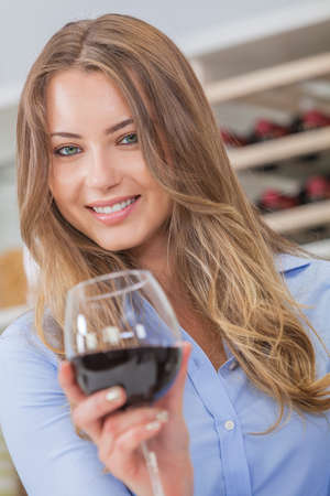 Beautiful young blond woman smiling and drinking a glass of red wine with bottles on a rack behind herの写真素材