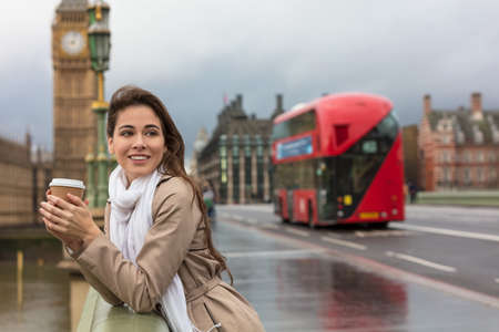 Girl or young woman drinking coffee in a disposable cup on Westminster Bridge with Big Ben and red double decker bus in the background, London, England, Great Britainの写真素材