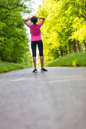 Beautiful mixed race African American young woman girl teenager fitness running jogging resting on road lined with spring or summer green treesの写真素材