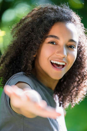 Outdoor portrait of beautiful happy mixed race African American girl teenager female young woman smiling with perfect teeth holding hand to cameraの写真素材