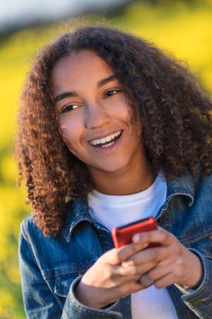 Outdoor portrait of beautiful happy mixed race African American girl teenager female young woman smiling with perfect teeth sending text on cell phoneの写真素材