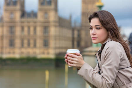 Beautiful sad, depressed or thoughtful young woman in London on Westminster Bridge over the River Thames drinking takeout coffee by Big Benの写真素材