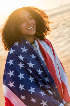 Outdoor portrait of beautiful happy mixed race African American girl teenager female young woman wrapped in the stars and stripes US flag on a beach smiling laughing with perfect teeth in golden sunset evening sunshineの写真素材