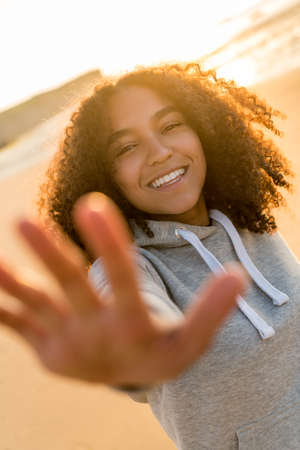 Outdoor portrait of beautiful happy mixed race African American girl teenager female young woman on a beach reaching out to camera, smiling and laughing with perfect teeth in golden sunset evening sunshineの写真素材