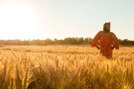 African woman in traditional clothes standing with her hands on her hips in field of barley or wheat crops at sunset or sunriseの写真素材