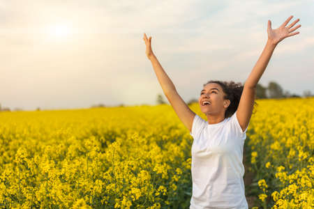 Outdoor portrait in golden evening sunshine of beautiful happy mixed race African American girl teenager female young woman smiling laughing arms raised celebrating in field of yellow flowersの写真素材