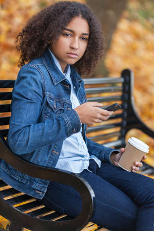 Beautiful mixed race African American girl teenager female young woman using cell phone and drinking takeaway coffee outside sitting on a park bench in autumn or fall looking sad depressed or thoughtfulの写真素材
