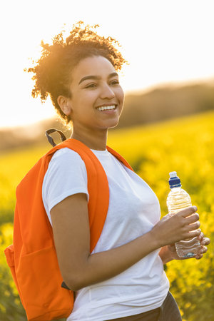Outdoor portrait in golden evening sunshine of beautiful happy mixed race African American girl teenager female young woman smiling laughing with perfect teeth hiking with orange rucksack and water bottle in field of yellow flowersの写真素材