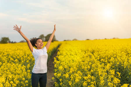 Outdoor portrait in golden evening sunshine of beautiful happy mixed race African American girl teenager female young woman smiling laughing arms raised celebrating in field of yellow flowersの写真素材