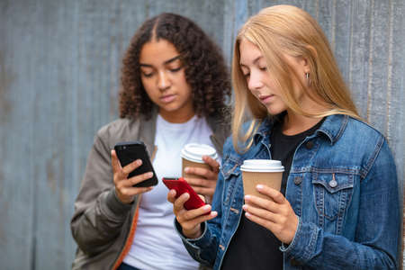 Interracial friends pretty blonde teenager and mixed race African American girl female young woman using their smart phones for social media and drinking takeout coffee outsideの写真素材
