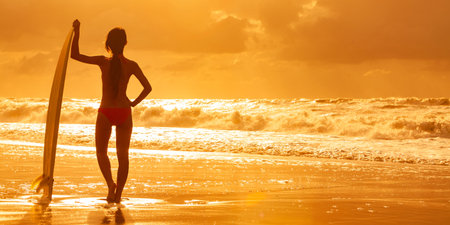 Rear view of beautiful sexy young woman surfer girl in bikini with white surfboard on a beach at...