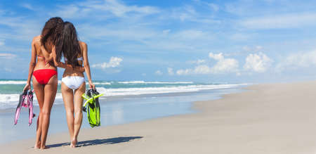 Panoramic banner rear view of two beautiful young women in red and white bikinis with snorkel, mask & flippers embracing on a deserted beach with blue skyの写真素材